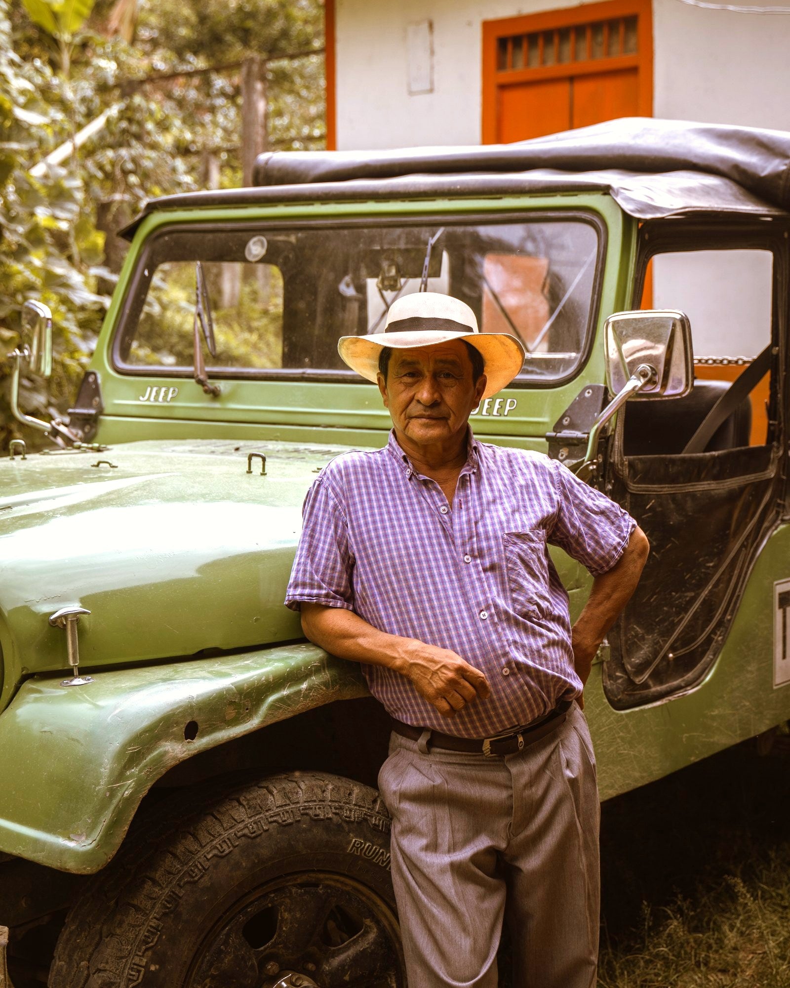 León Campo mit Hut und kariertem Hemd, angelehnt an seinen grünen Willys-Jeep zwischen den Kaffeebäumen